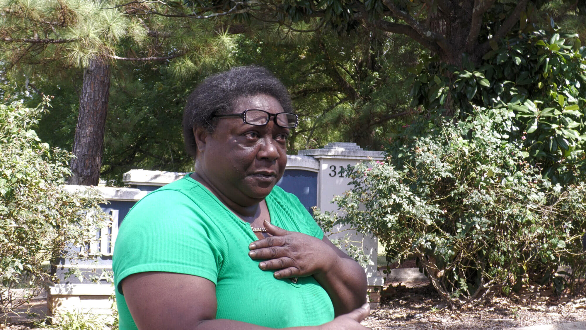 A woman in a green shirt with glasses resting on her head holds one hand to her chest while standing outside.