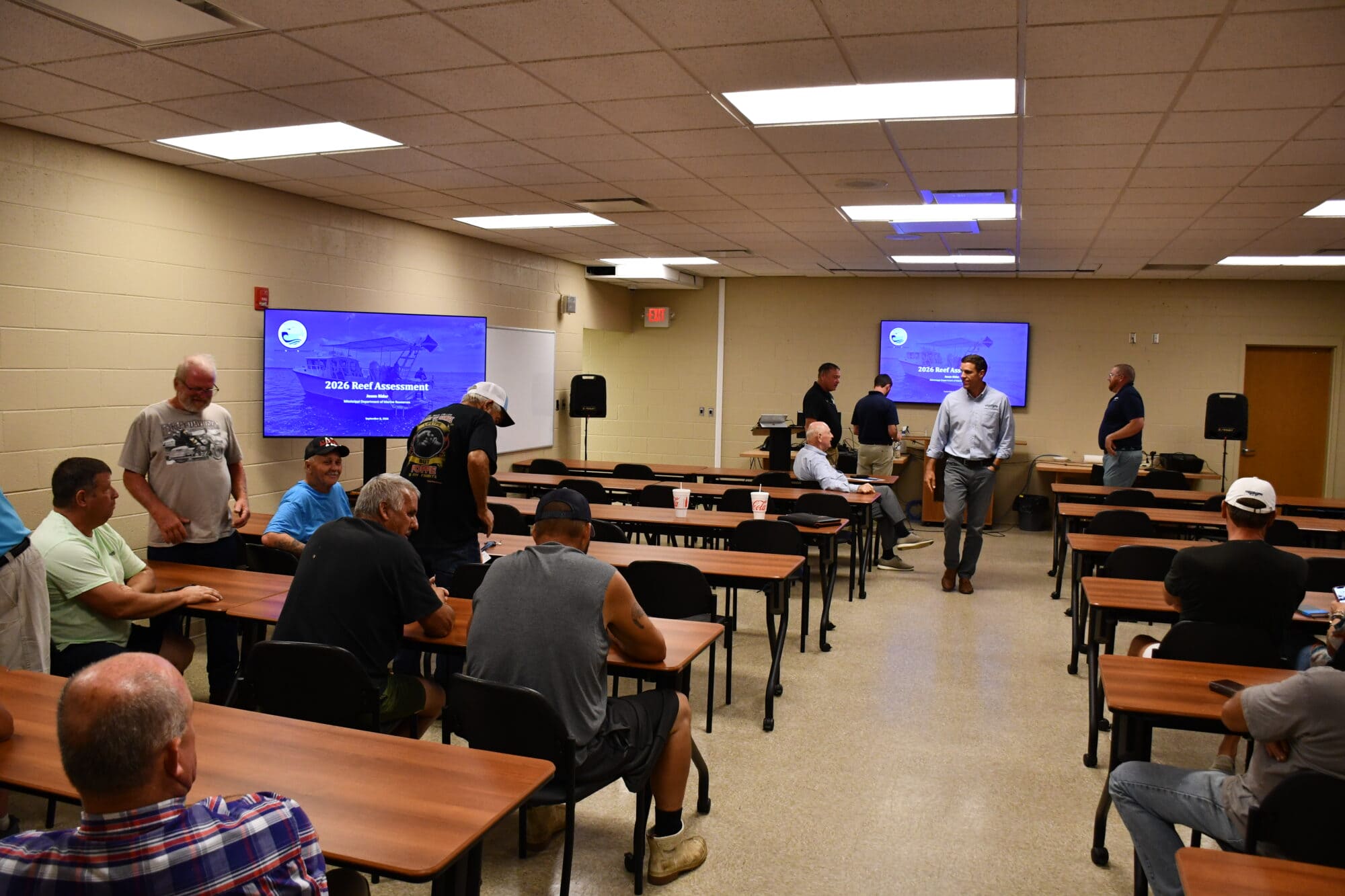 Fishermen and state marine resources officials gather in a meeting room as screens display a presentation titled “2026 Reef Assessment.”