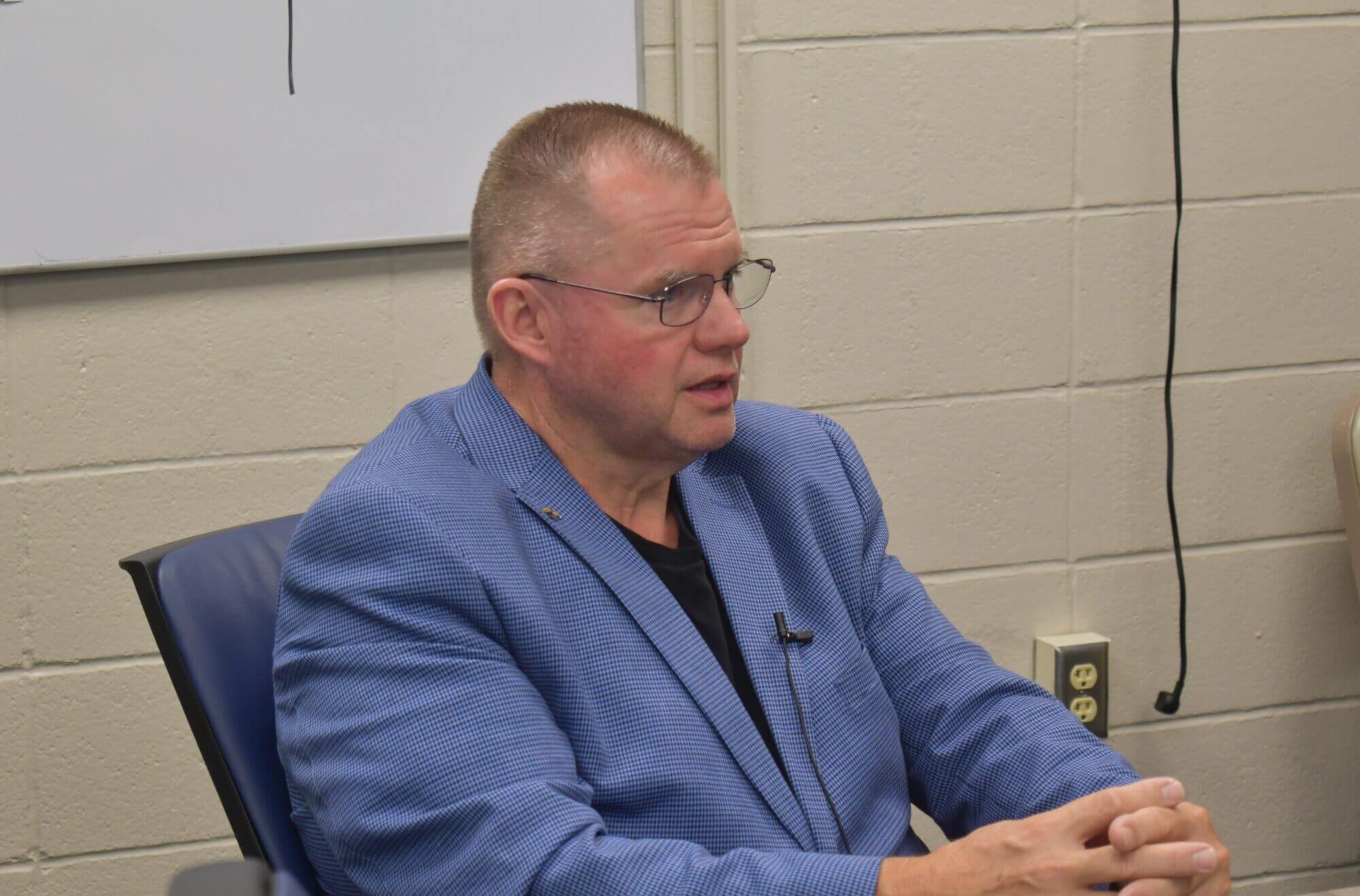 A man wearing a blue blazer over a black shirt sits at a table.