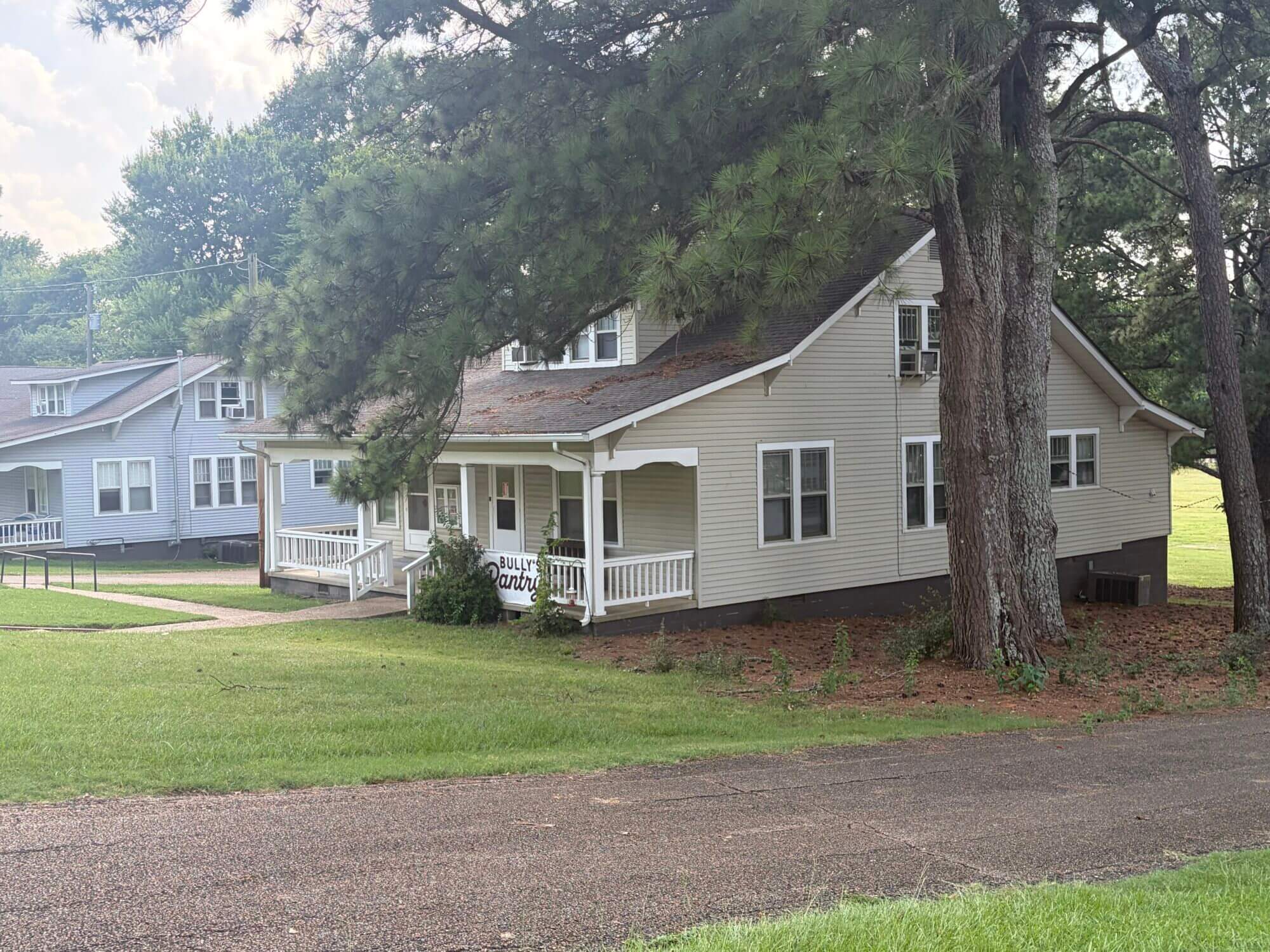 A tan house with a dark roof and white front porch sits behind several large pine trees on a grassy Mississippi State University campus.
