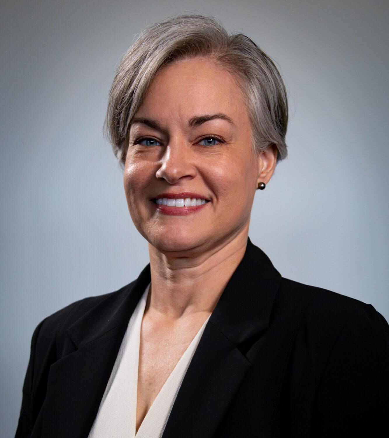 A woman wearing a white blouse and black blazer smiles in front of a light gray background.