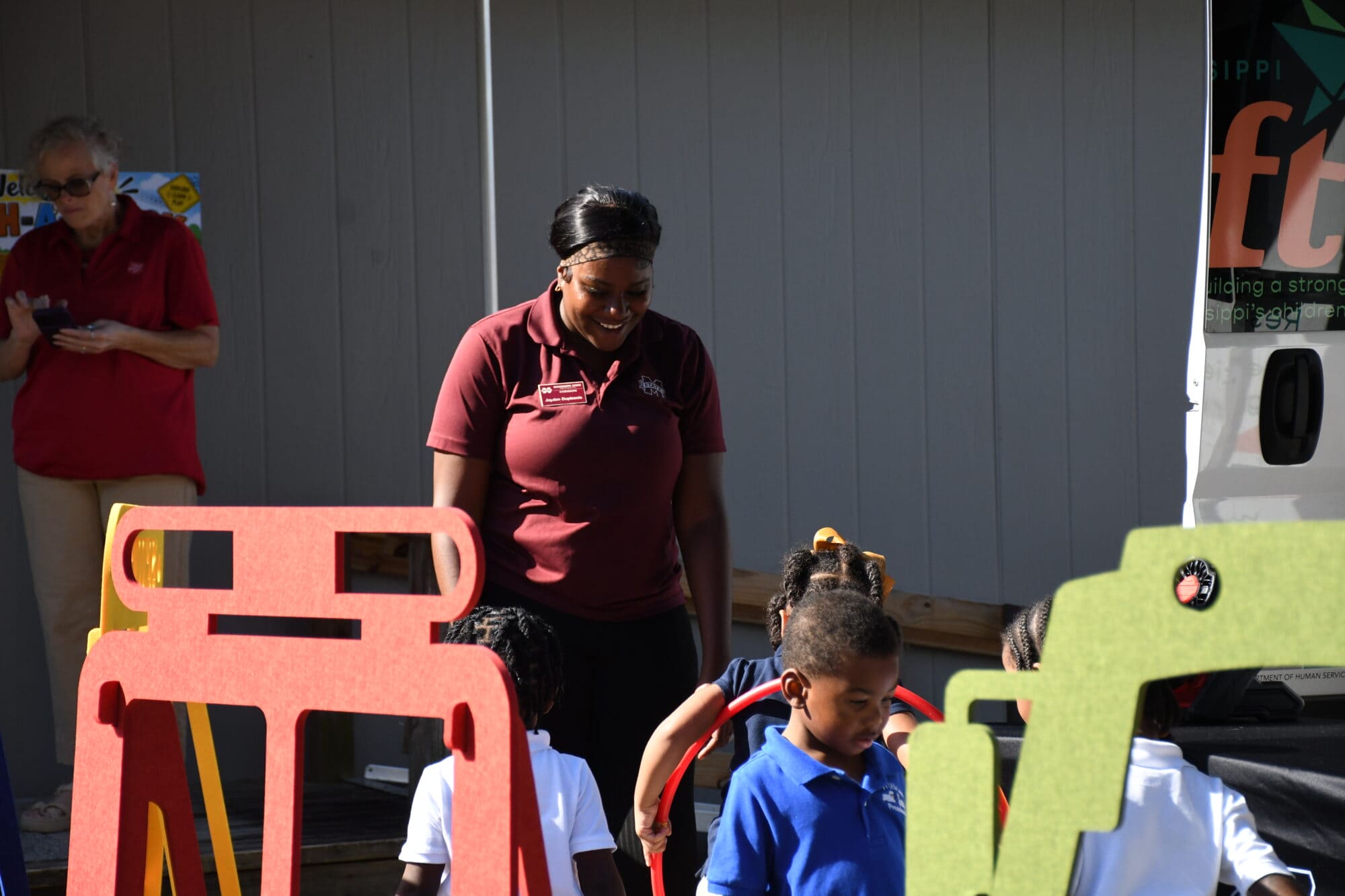 MS LIFT program associate Jayden Duplessis smiles while standing among children playing with colorful activity equipment at an outdoor event.