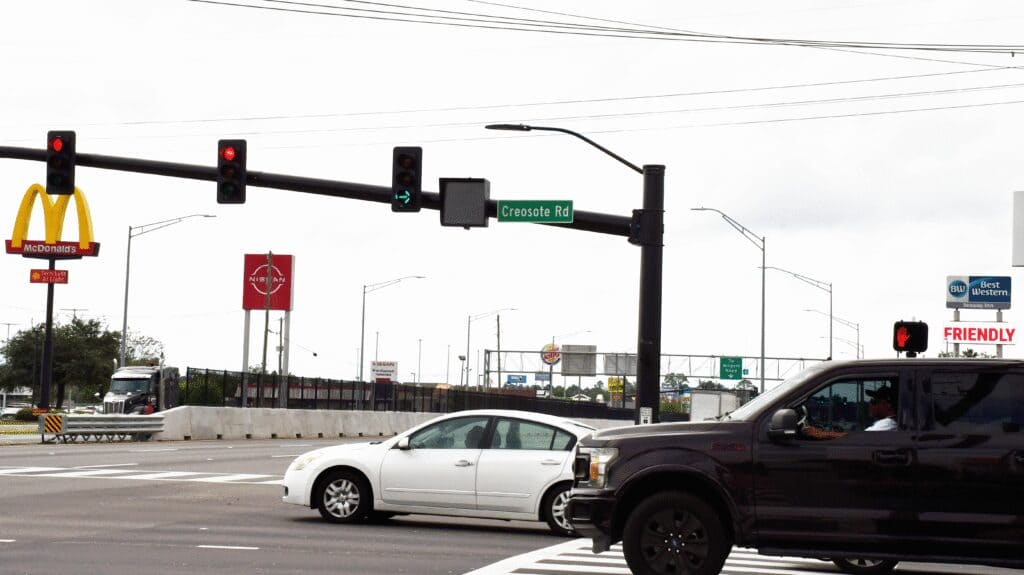 A white sedan and dark pickup truck travel through a busy intersection in Gulfport beneath traffic lights and a Creosote Road sign.