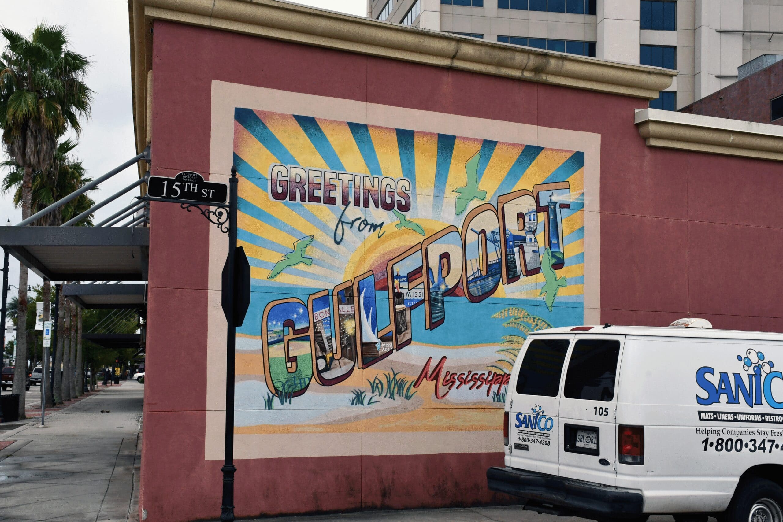 A colorful “Greetings from Gulfport, Mississippi” mural covers the side of a downtown building. A white service van is parked in front of the mural.