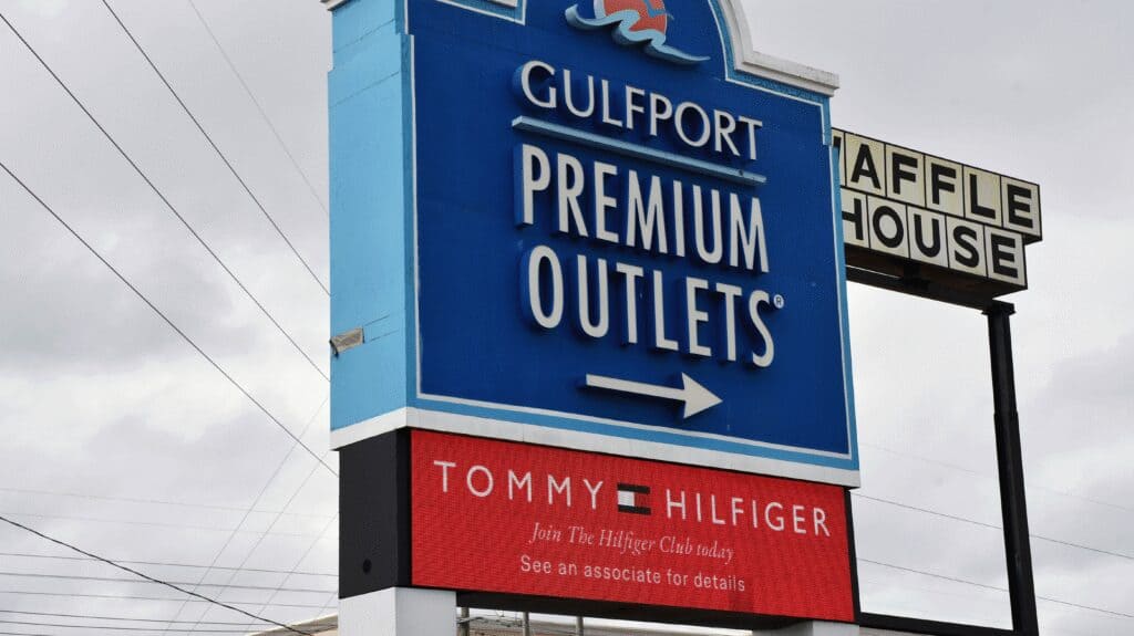 A large blue Gulfport Premium Outlets sign with a white directional arrow stands next to a Waffle House sign beneath an overcast sky.