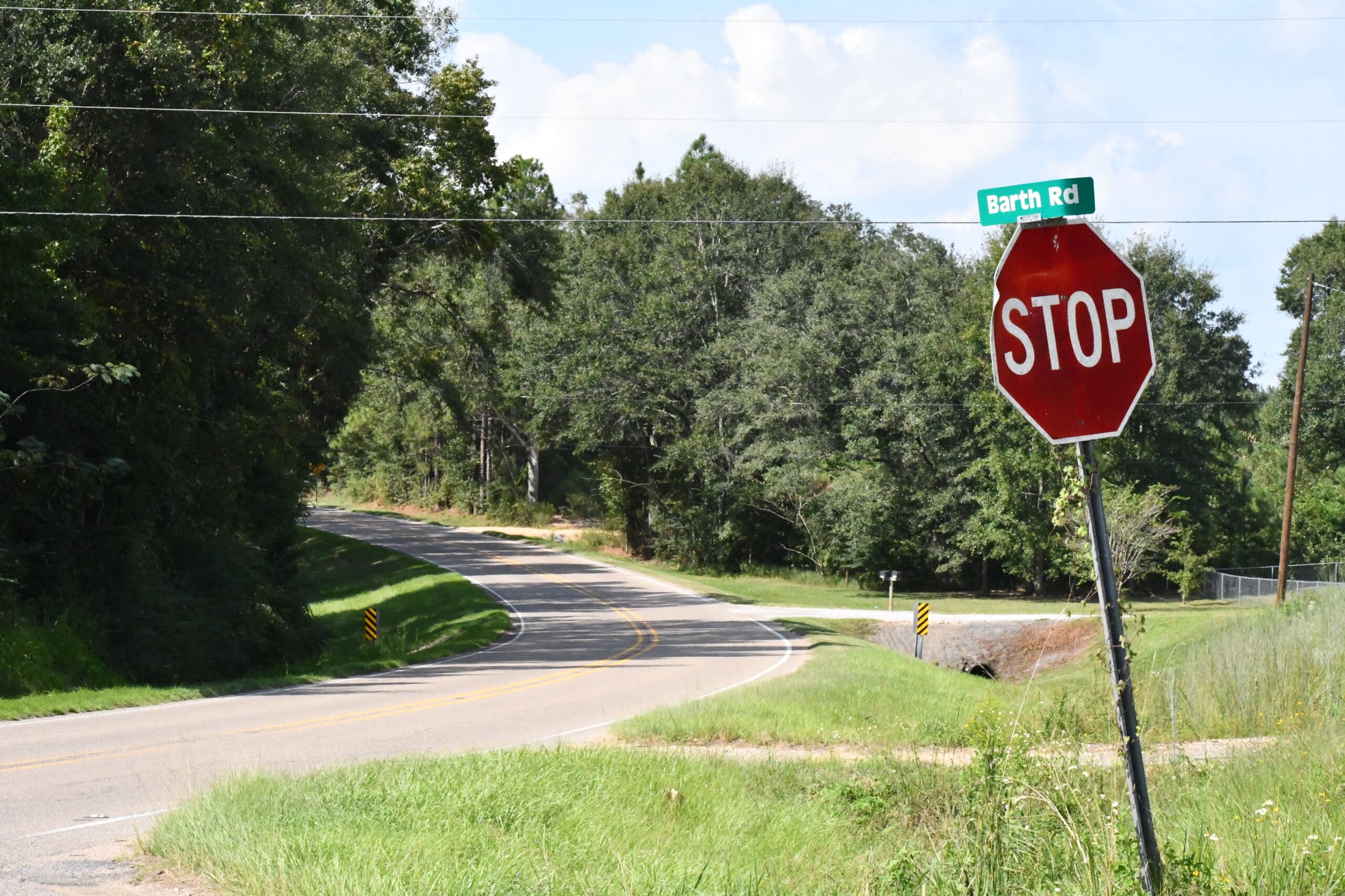A stop sign and Barth Road street sign stand beside a two-lane road surrounded by trees and grass.