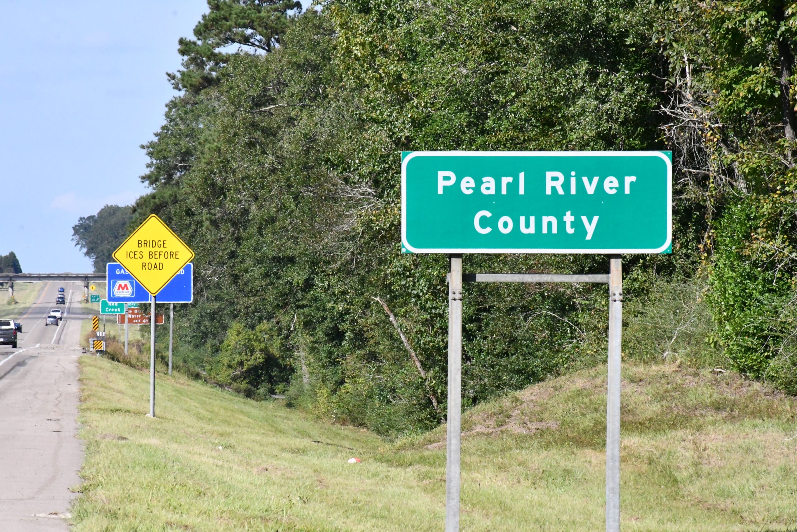 A green Pearl River County sign stands beside a highway lined with trees, with traffic and other road signs visible in the distance.