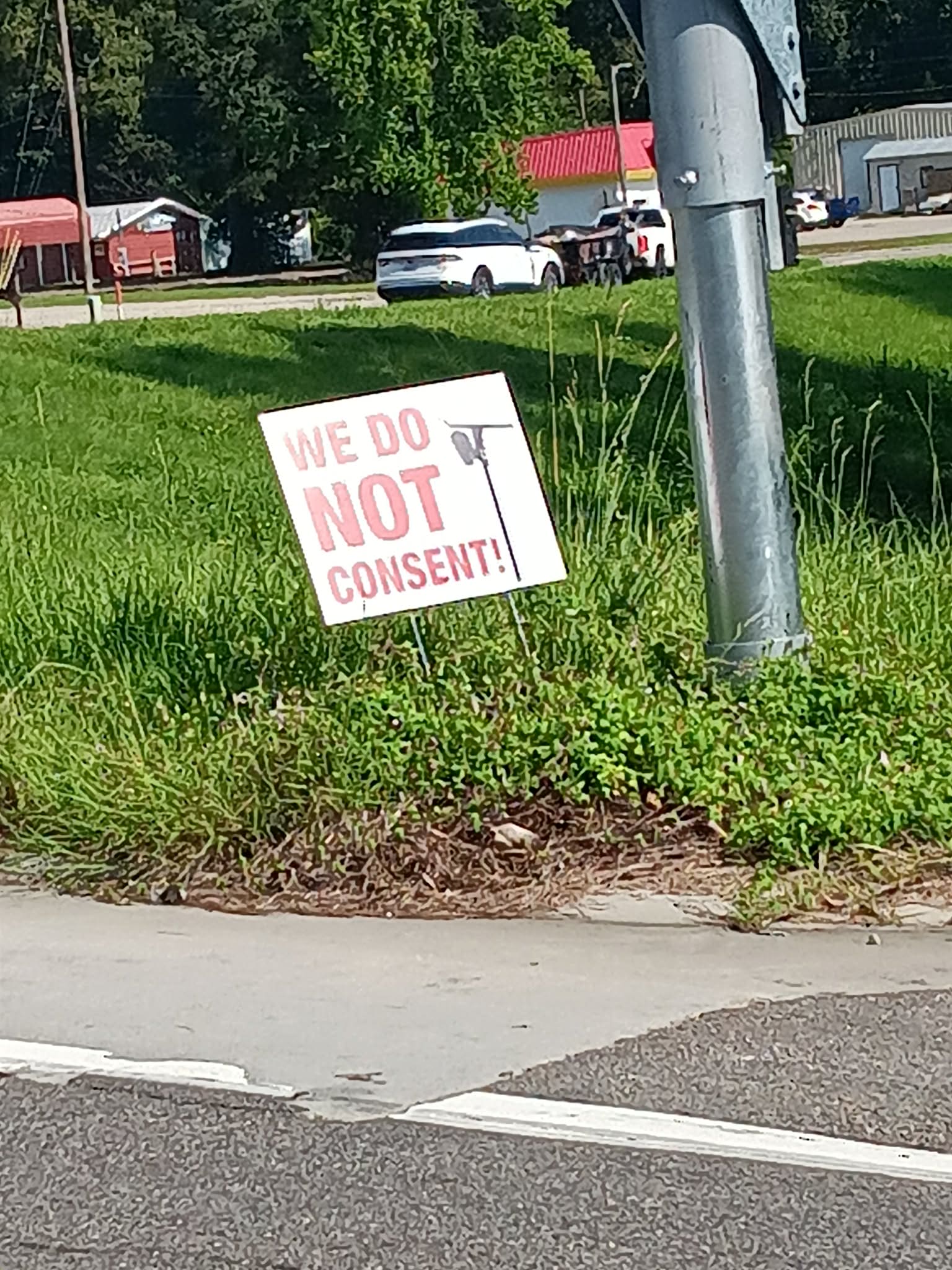 A white yard sign reading “WE DO NOT CONSENT!” stands in grass near a Picayune intersection.
