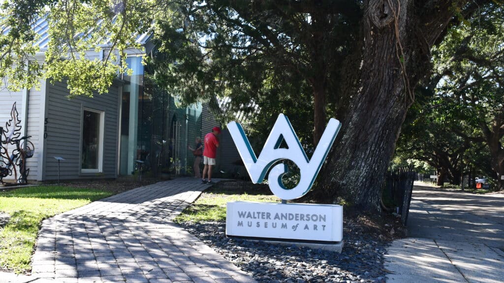 A white Walter Anderson Museum of Art sign stands beneath a large tree beside a paved walkway leading to the museum's entrance.