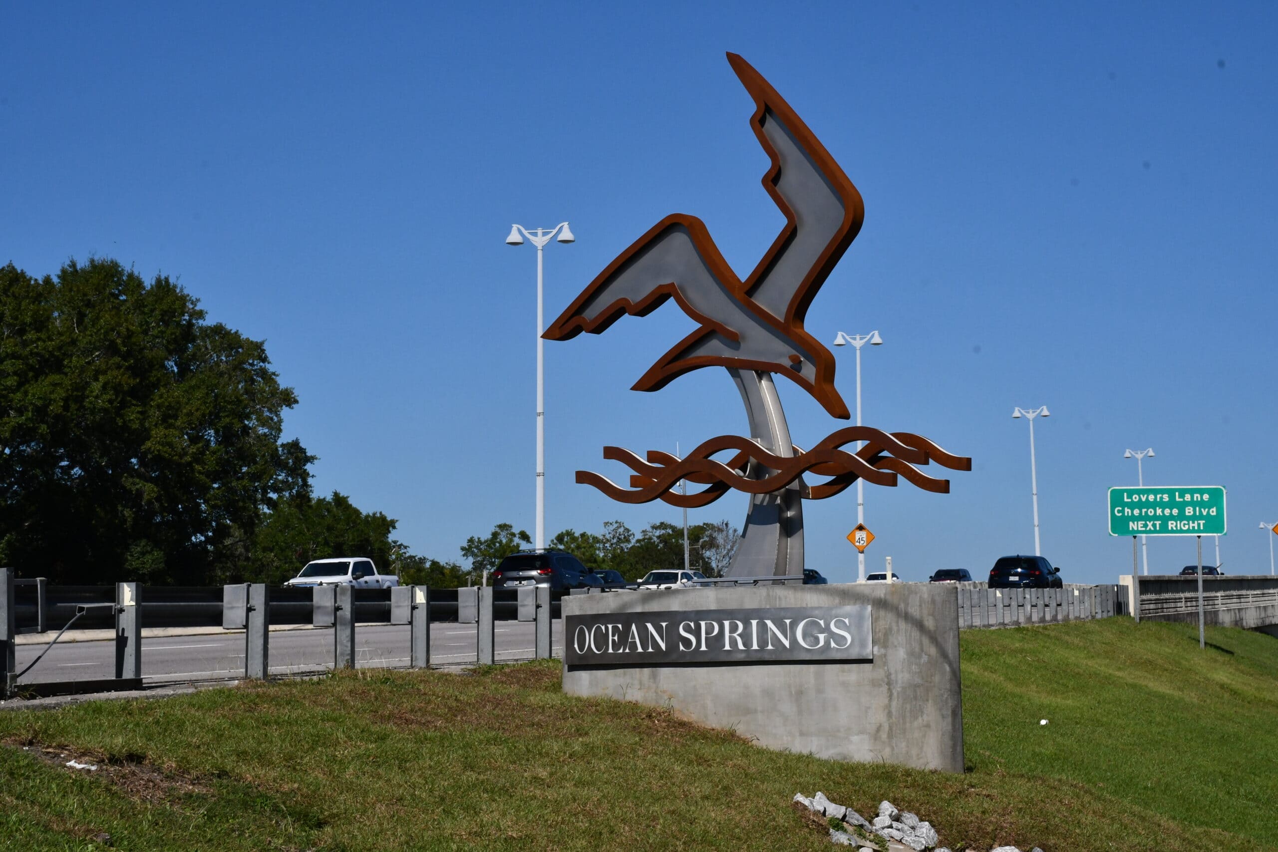 A large metal sculpture of a bird in flight rises above wave-shaped metal elements near a bridge in Ocean Springs. The concrete base reads "Ocean Springs."